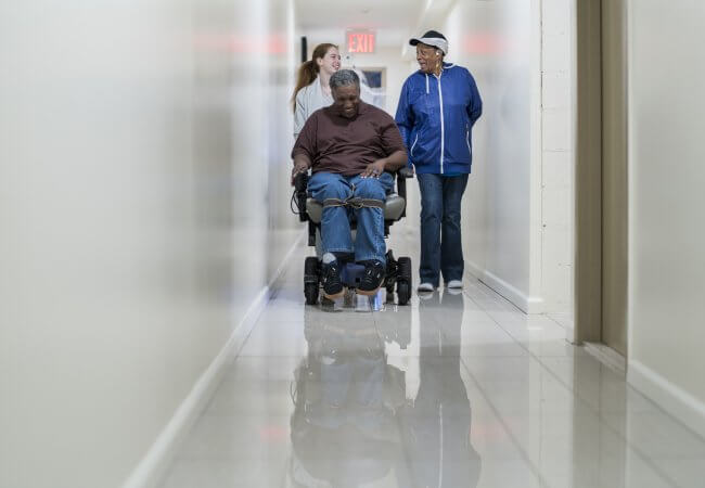 The disabled paralozed African-American man in wheelchair, accompanied with Black woman and a nurse in a corridor.